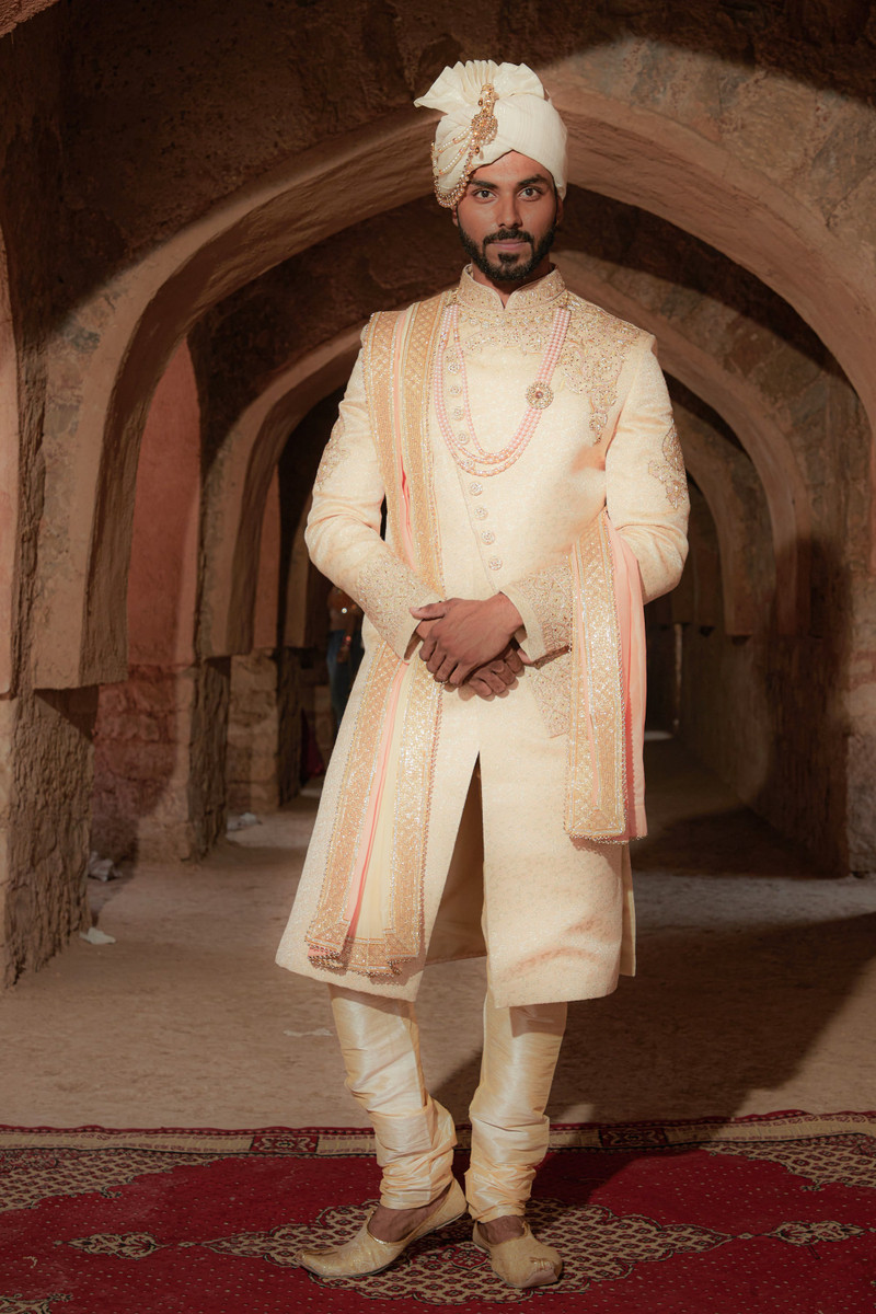 An ivory sherwani with a safa in a sandstone corridor.